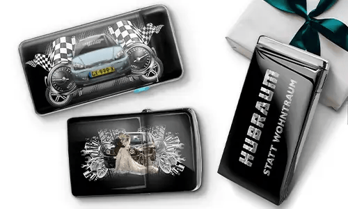 Drei schwarze Geldbörsen mit Auto-Designs und karierten Flaggen, vor einem weißen Hintergrund. Eine zeigt ein Auto mit Rädern, eine andere ein Paar, und eine trägt den Text „Hubraum statt Wohntraum“.